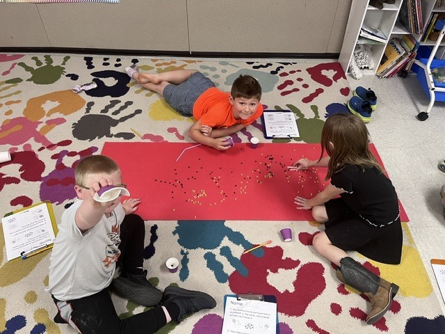 Three elementary-aged children are working on a colorful, handprint-patterned rug around a long red mat. The mat has small black beans and yellow macaroni pieces scattered across it. One boy lies on his stomach at the top of the mat, a girl sits on the right side manipulating the pieces, and a boy in the bottom left foreground looks at the camera while holding up a small purple paper cup.  image