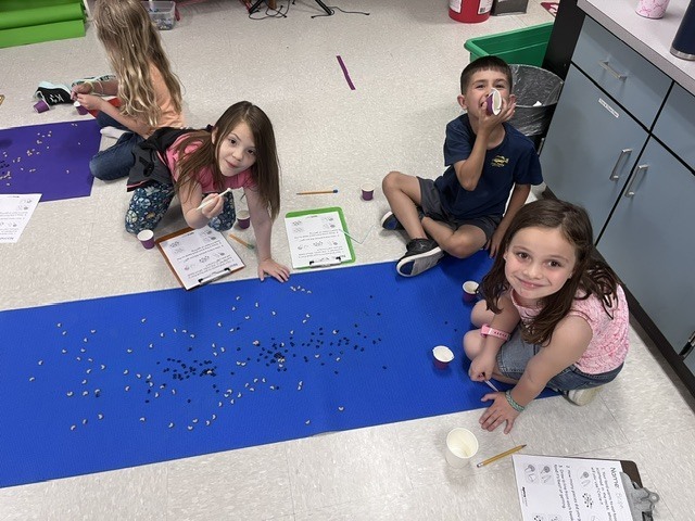  A group of four children are gathered on the classroom floor around a long, bright blue mat scattered with small black and tan pieces. A young girl in the foreground on the right smiles directly at the camera, while another girl on the left leans forward holding a straw like a tool. Behind them, a boy sits cross-legged holding a small cup up to his face, and worksheets with clipboards are scattered around the workspace.