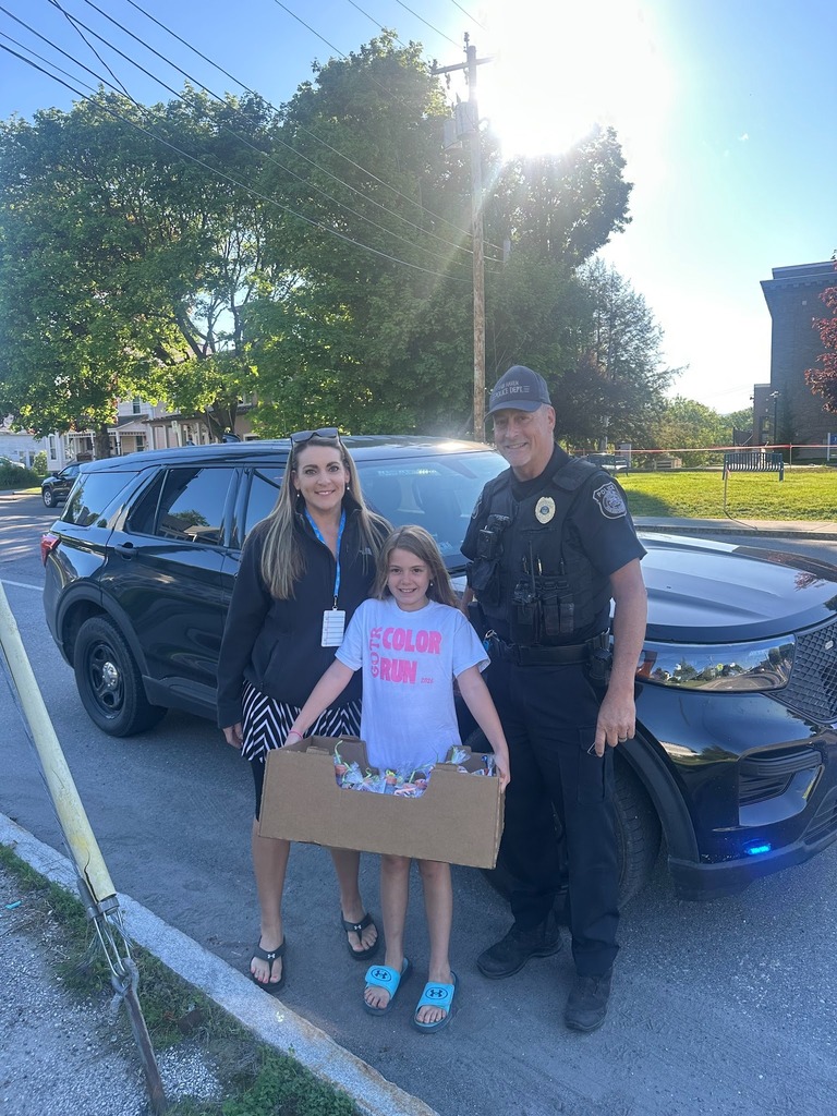 A Girls on the Run student stands between an adult woman and a Fair Haven police officer in front of a police SUV on a sunny day. The student holds a box filled with care packages and treats prepared for the Fair Haven Police and Fire Departments as part of a community service project.