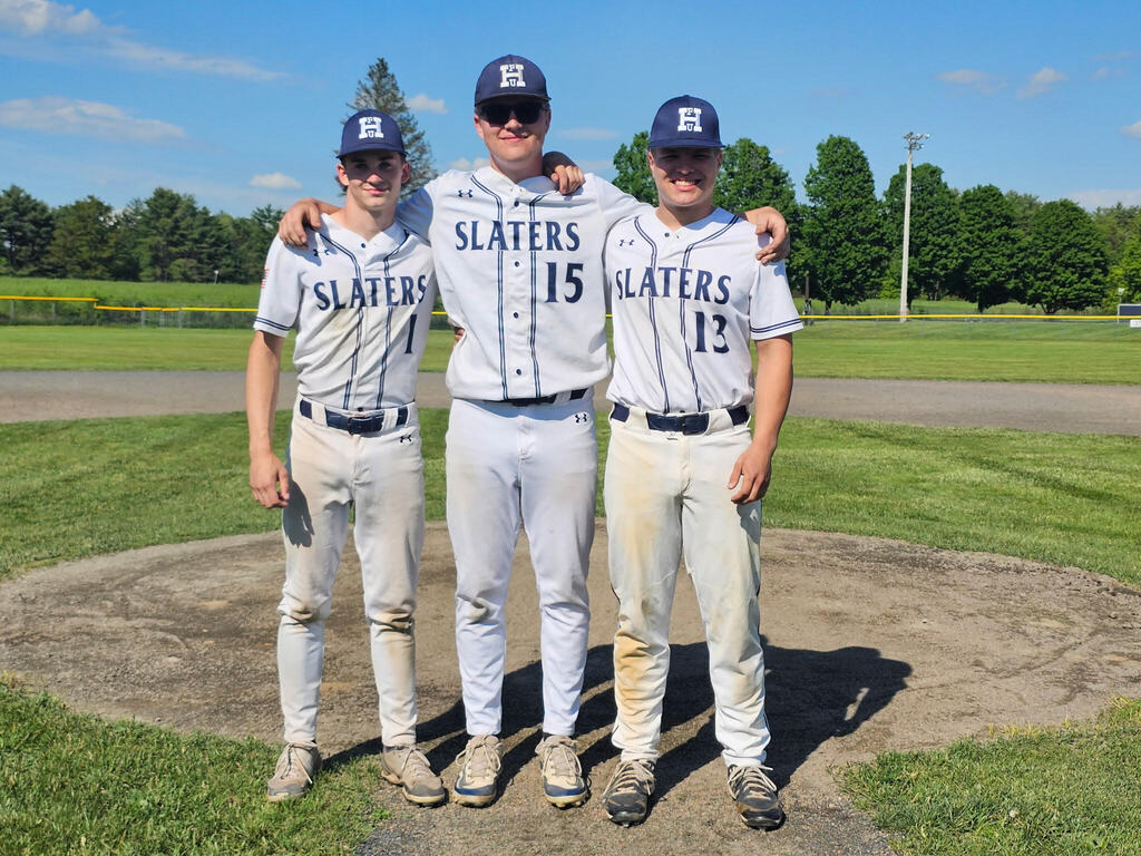 Three Fair Haven Union High School senior baseball players stand together on the pitcher’s mound with their arms around each other during Senior Night. The players wear gray Slaters baseball uniforms and navy caps, with a sunny field and trees in the background.