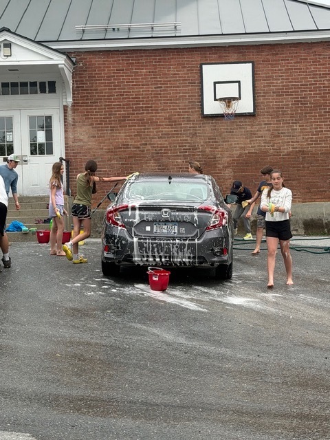 Students wash a black sedan covered in soap in a school parking lot. One student sprays water while others scrub the car with sponges and buckets nearby.