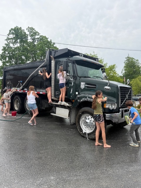 Several children wash a large black dump truck outdoors on a cloudy day, using sponges, soap, and hoses. Soap suds cover parts of the truck as students work together around it.