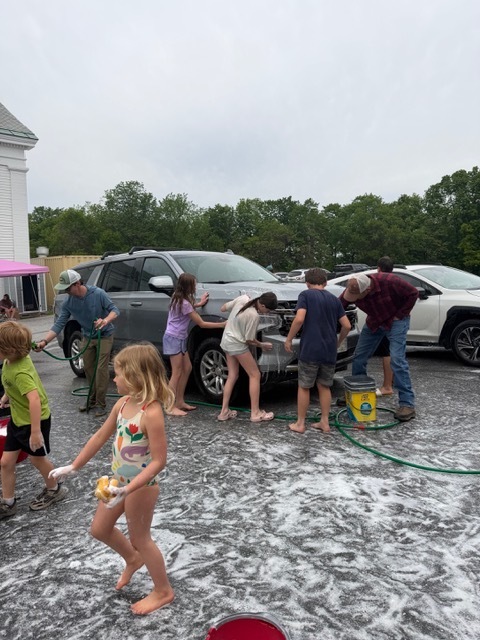 Children and adults wash a gray SUV in a parking lot covered in soap suds. Students use sponges and hoses while others walk nearby carrying cleaning supplies.