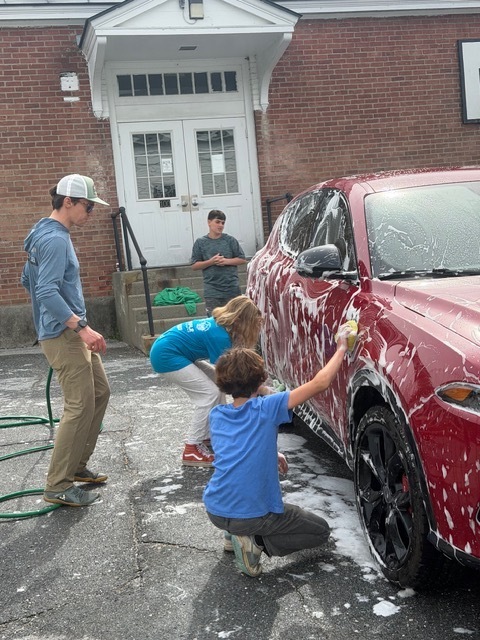 Children and an adult work together washing a red SUV covered in thick soap suds. Two students scrub the side of the vehicle while others stand nearby with a hose and cleaning supplies.