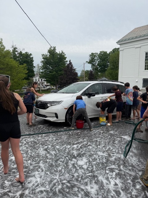 A group of students washes a white minivan during a community car wash event. Children scrub the vehicle with soapy sponges while buckets and hoses sit nearby.