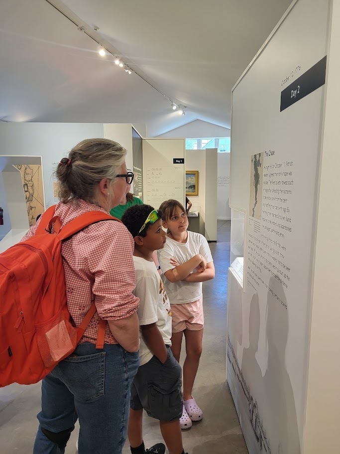 An adult and two children read an informational museum display together inside a history exhibit with white walls and educational panels.