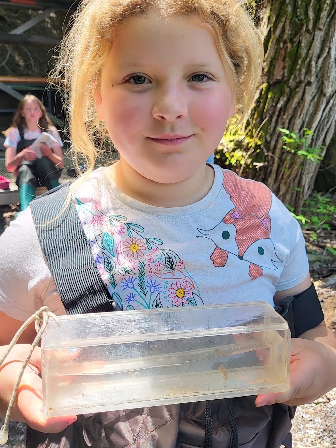 A smiling young girl wearing waders holds a clear observation container with small aquatic creatures inside while standing in a wooded outdoor classroom area.