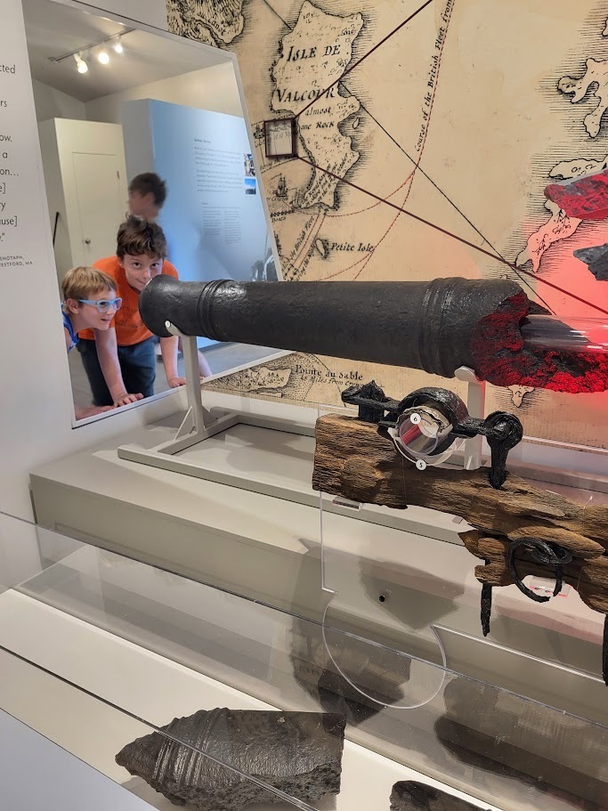 Two children peer through a display opening at a historic cannon exhibit inside a museum featuring maps and artifacts related to Lake Champlain history.