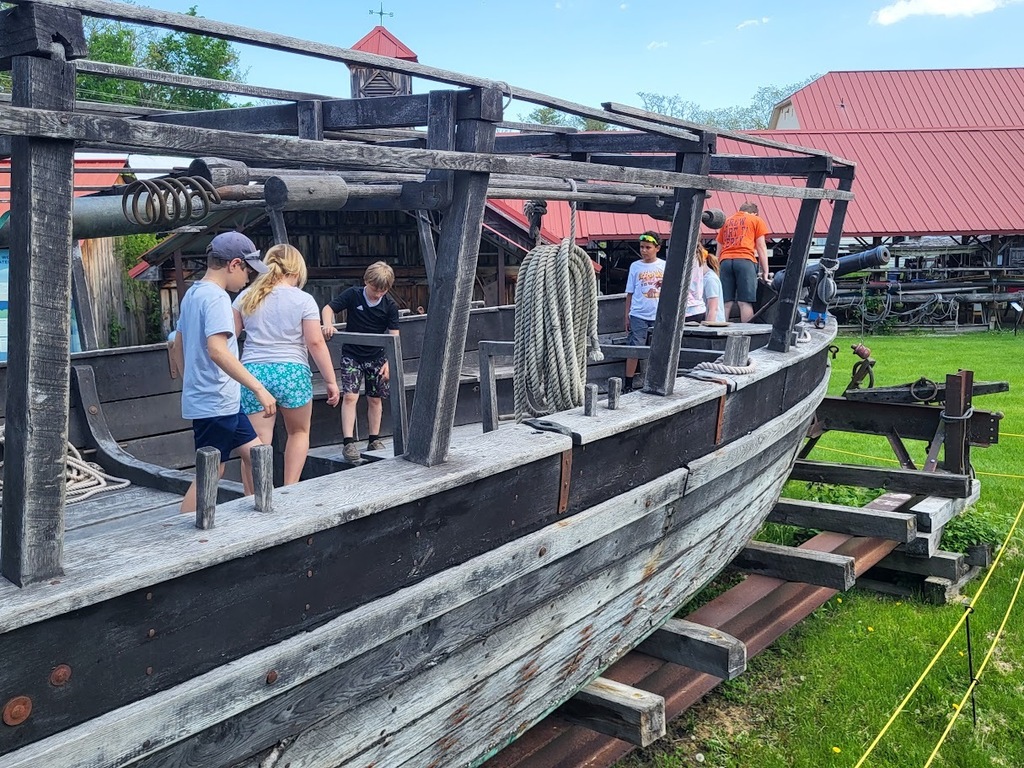 Children explore a large wooden historic ship replica outdoors at a museum site with red-roofed buildings in the background.