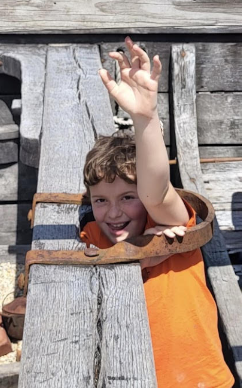 A smiling child in an orange shirt playfully peeks through a wooden restraint on a historic ship exhibit outdoors.