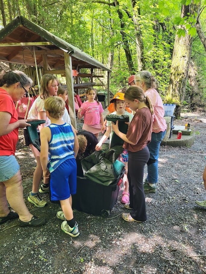 A group of children and adults gather outdoors in a wooded area around large storage bins and waders, preparing for an environmental education activity near the water.