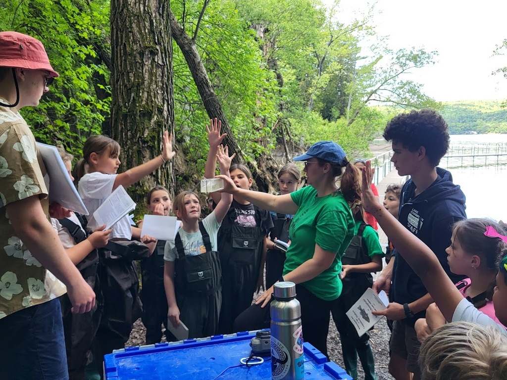 An instructor in a green shirt holds up a clear container while students in waders raise their hands during an outdoor science lesson beside a lake.