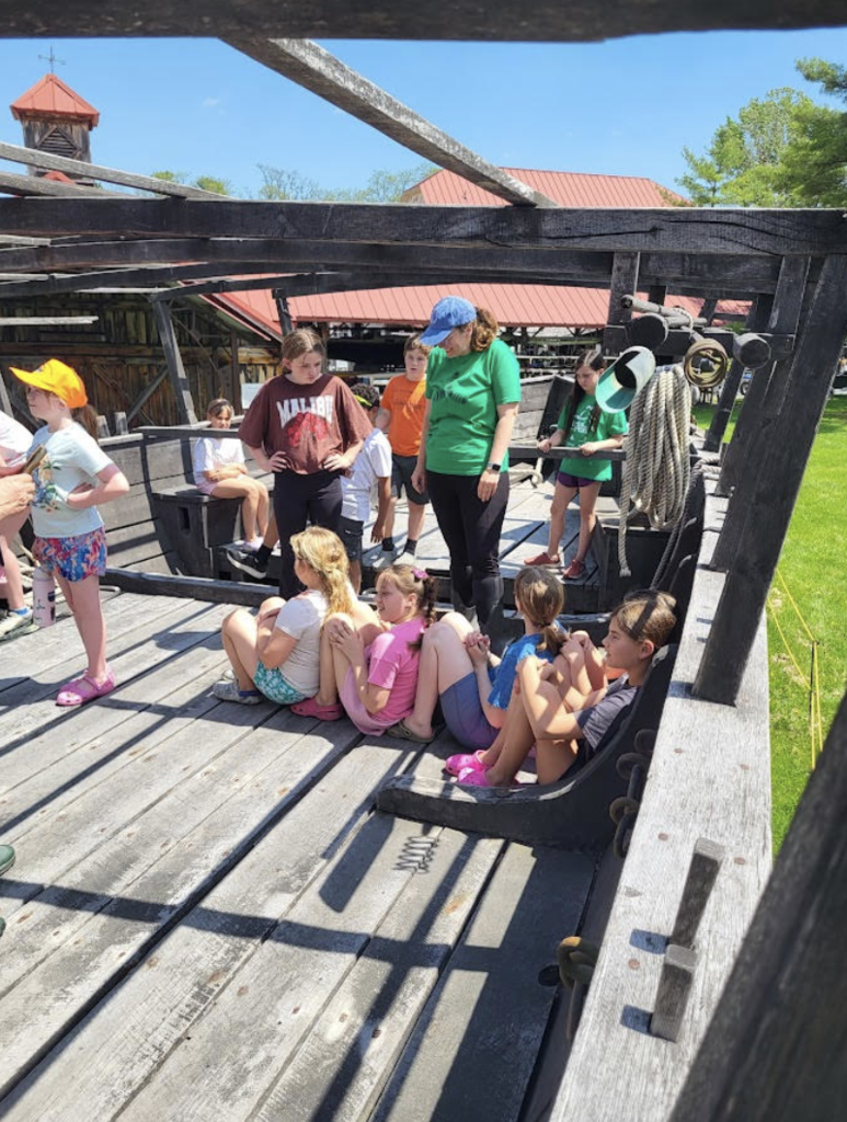 Students and an instructor sit and stand aboard a large wooden ship replica during an outdoor educational tour on a sunny day.