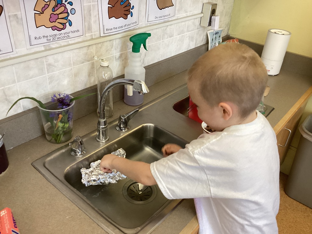 A kindergarten student places pennies into a handmade aluminum foil boat floating in a classroom sink during a STEM math activity. Handwashing posters hang above the sink.
