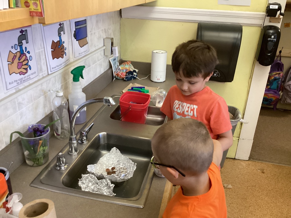 Two kindergarten students stand at a classroom sink testing an aluminum foil boat filled with pennies to see how much weight it can hold before sinking.