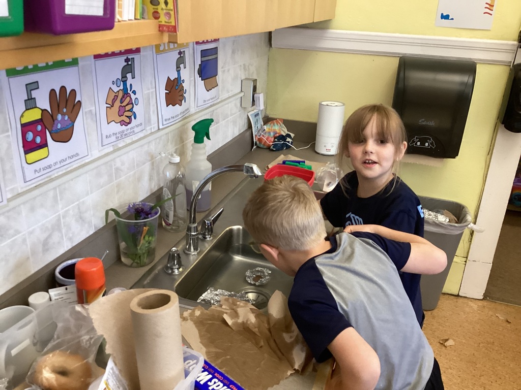 Two kindergarten students work together at a classroom sink during a STEM activity involving aluminum foil boats and pennies while discussing their results.