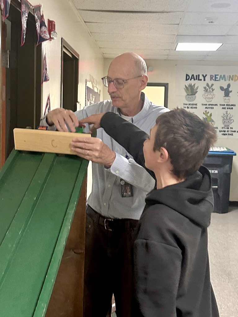 A staff member and student place toy cars at the top of a large green ramp as part of a hands-on STEM activity in a school hallway.