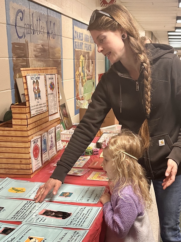 A parent and young child look at student writing and artwork displayed on a table during the Celebration of Learning event.