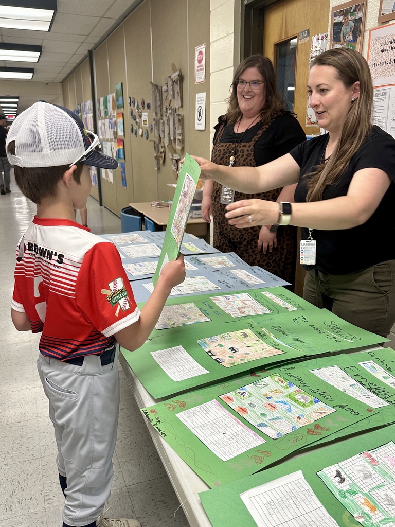A student in a baseball uniform presents a project display board to two teachers standing behind a table filled with student work during a school showcase event.