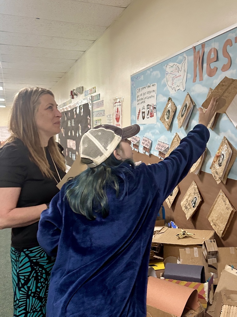 A parent and student examine a classroom display board featuring student-created projects and writing assignments mounted on brown paper.