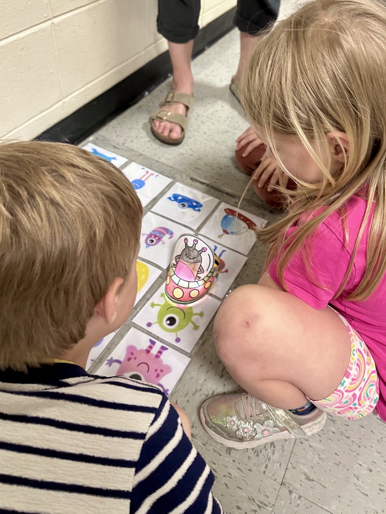 Two elementary students sit on the floor working together on a space-themed matching game with laminated cards featuring cartoon aliens and spaceships.