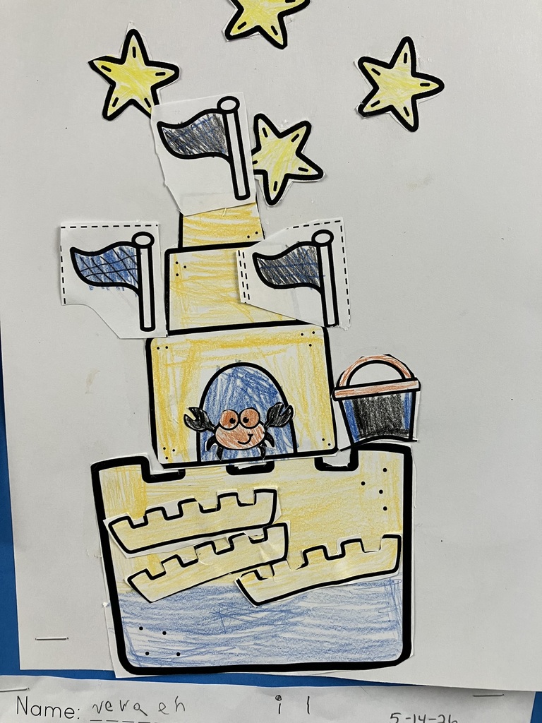  A vertical, close-up shot of a student's art project on white paper. The artwork is a paper cutout collage of a yellow sandcastle with various pieces layered on top of each other. A small cutout of a smiling orange crab looks out from a blue arched doorway in the middle tier. Three blue-and-white checkered flags are glued near the top of the towers, a black bucket sits on a ledge, and four yellow stars are glued in the white space above the castle. The bottom of the castle is colored with blue crayon to look like water.