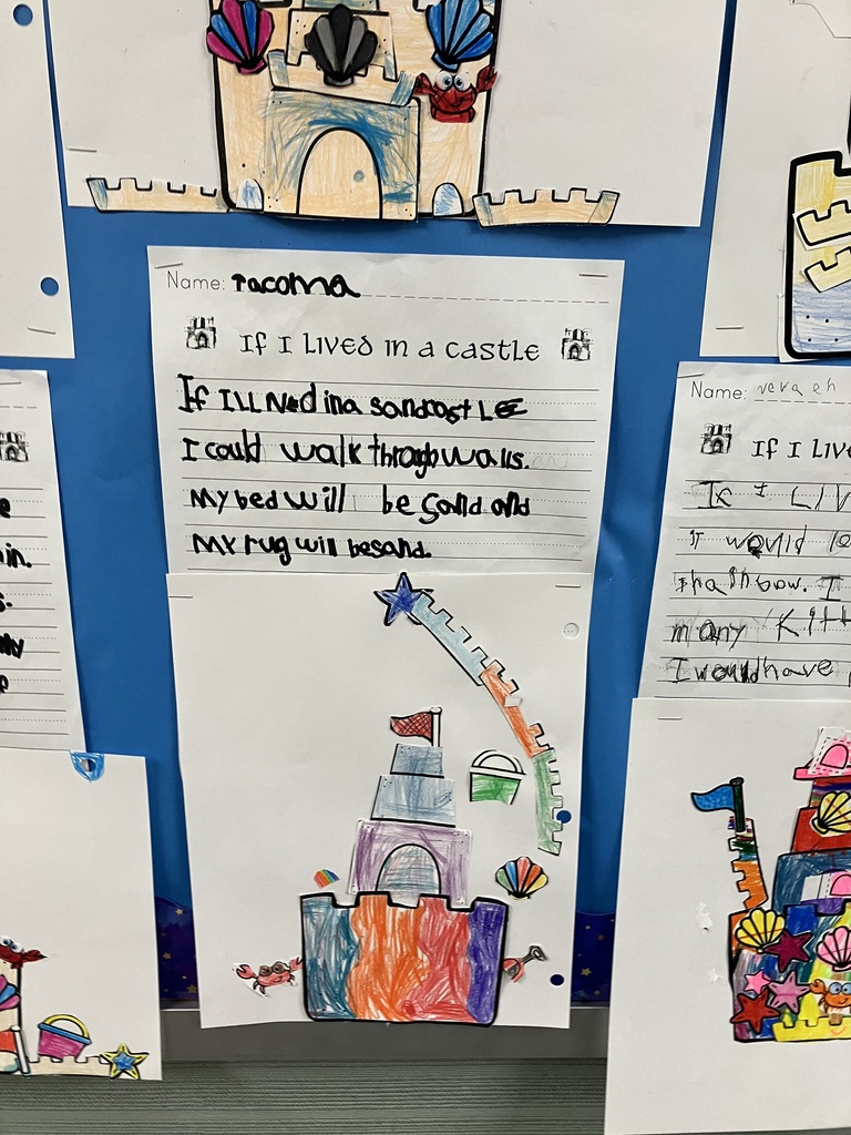 A close-up of a student project on a blue bulletin board. The handwritten text reads, "If I lived in a sandcastle I could walk through walls. My bed will be sand and my rug will be sand." Glued below the text is a creative paper collage of a castle. The castle base is vibrantly colored with vertical stripes of blue, orange, red, and purple crayon. A long, curved green and orange paper cutout, topped with a blue star, arches over the right side of the castle like a magical beam or ramp. 
