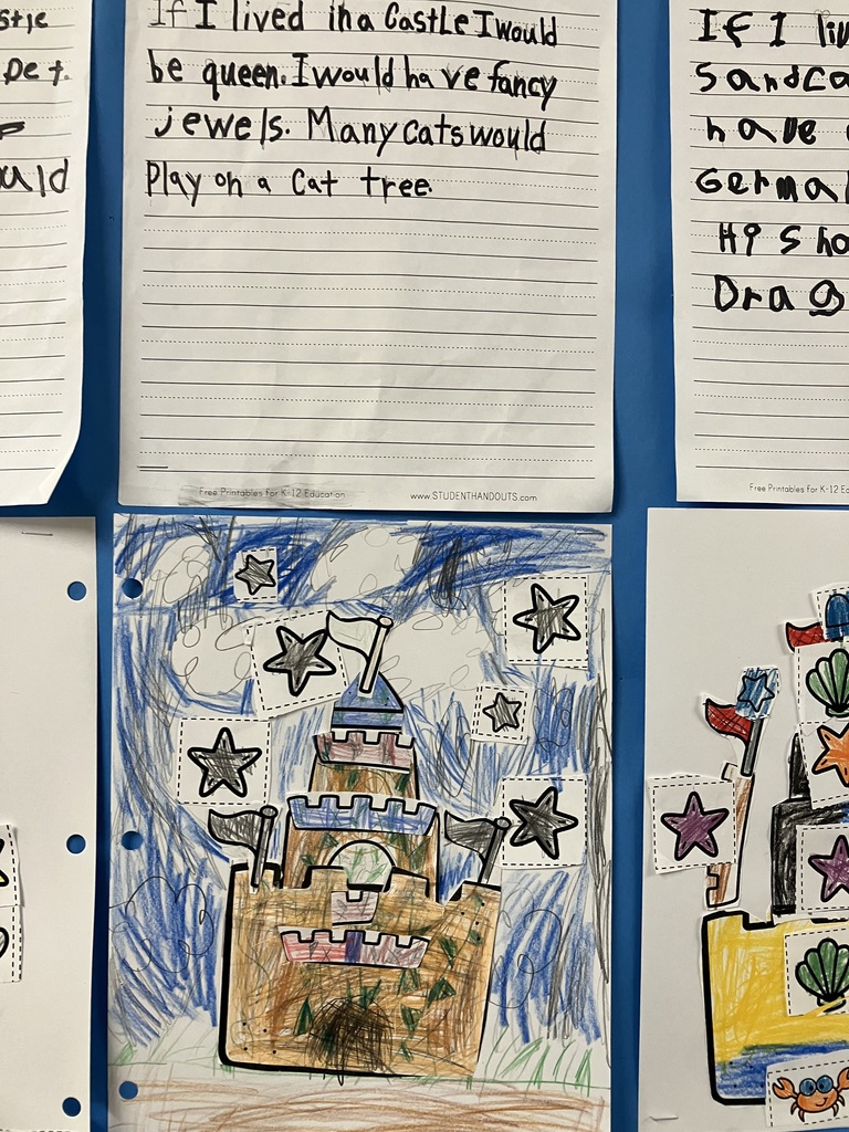  Alt Text: A close-up view of a first-grader's schoolwork on a blue background. The top page shows handwritten text by a studen reading, "If I lived in a castle I would be queen. I would have fancy jewels. Many cats would play on a cat tree." Below the writing is a drawing of a multi-tiered brown sandcastle heavily colored in with crayons. The background is filled with messy, dark blue and black scribbles representing a stormy or night sky, decorated with several cutout paper stars glued around the castle.