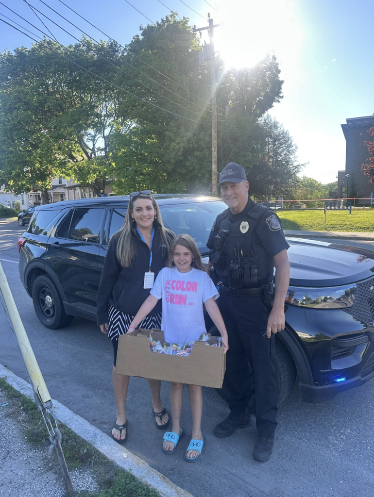 A student stands between a school staff member and a Fair Haven police officer while holding a box of colorful treat bags in front of a police cruiser on a sunny afternoon.
