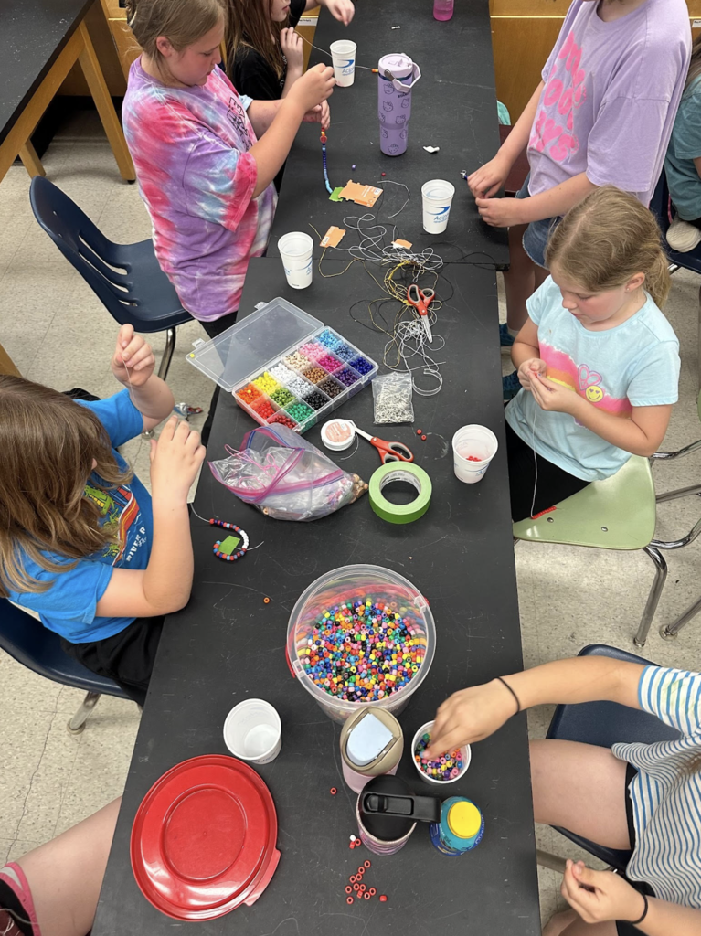 Students sit around a classroom table making colorful beaded bracelets, with containers of beads and craft supplies spread across the table.