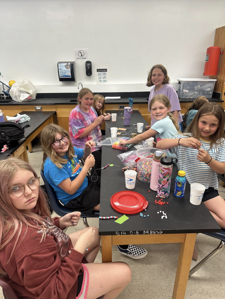 Several students smile while working on bracelet-making projects in a classroom filled with beads, string, and craft materials.