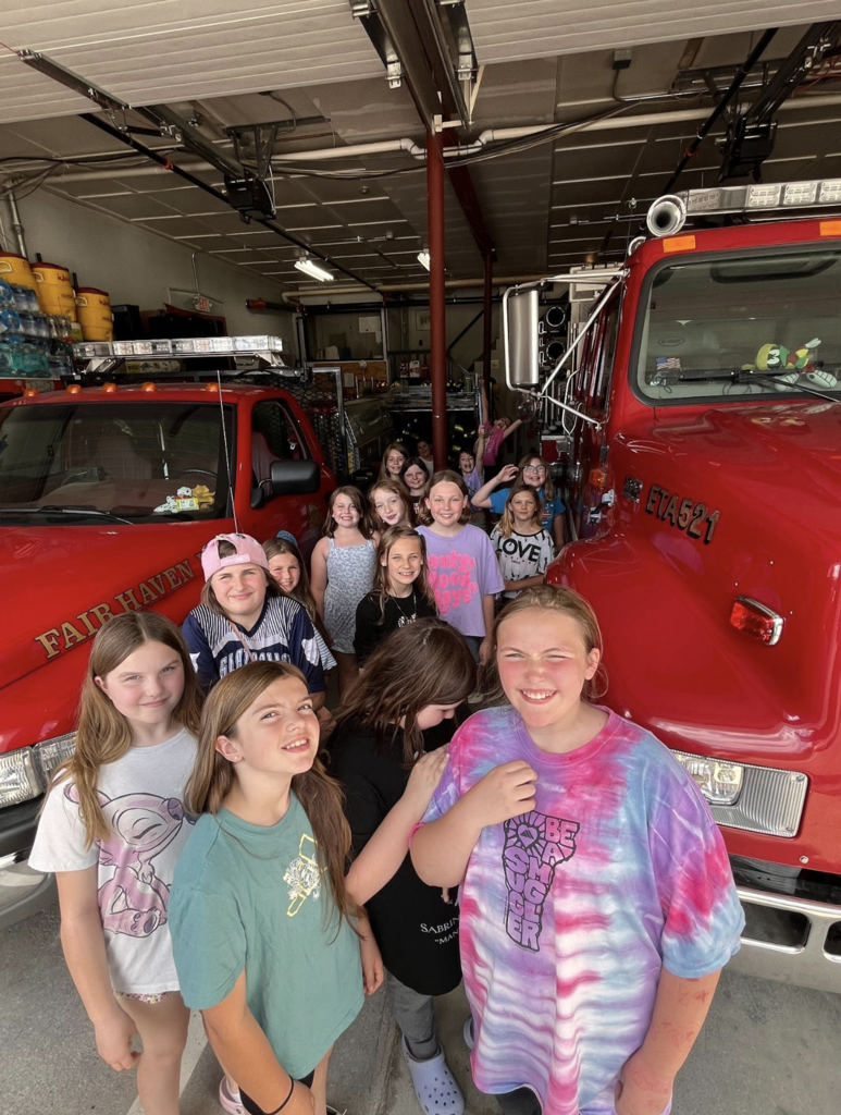 A large group of elementary students smiles for a photo inside the Fair Haven Fire Department garage between two bright red emergency vehicles.