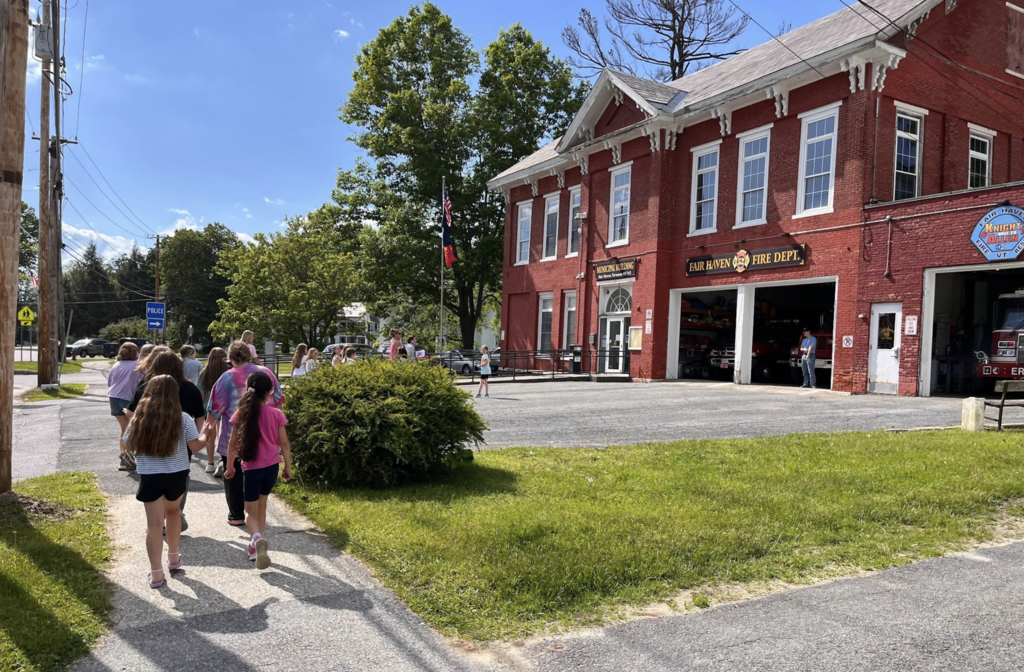 A group of elementary students walks toward the historic red-brick Fair Haven Fire Department building on a sunny day.