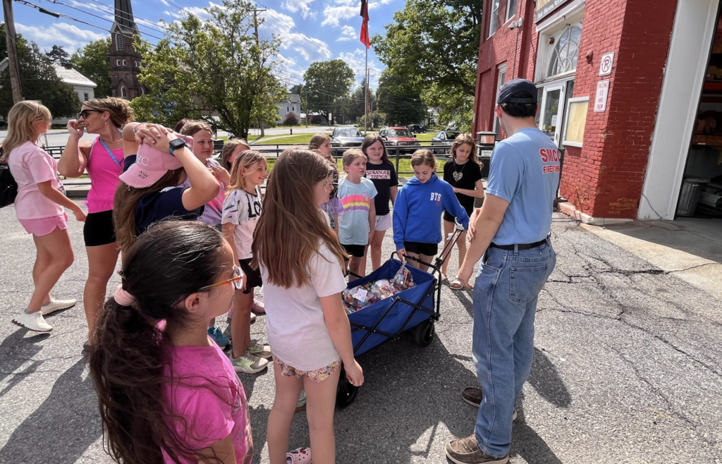 Students listen as a firefighter speaks outside the Fair Haven Fire Department, standing around a wagon filled with donated snacks and treats.