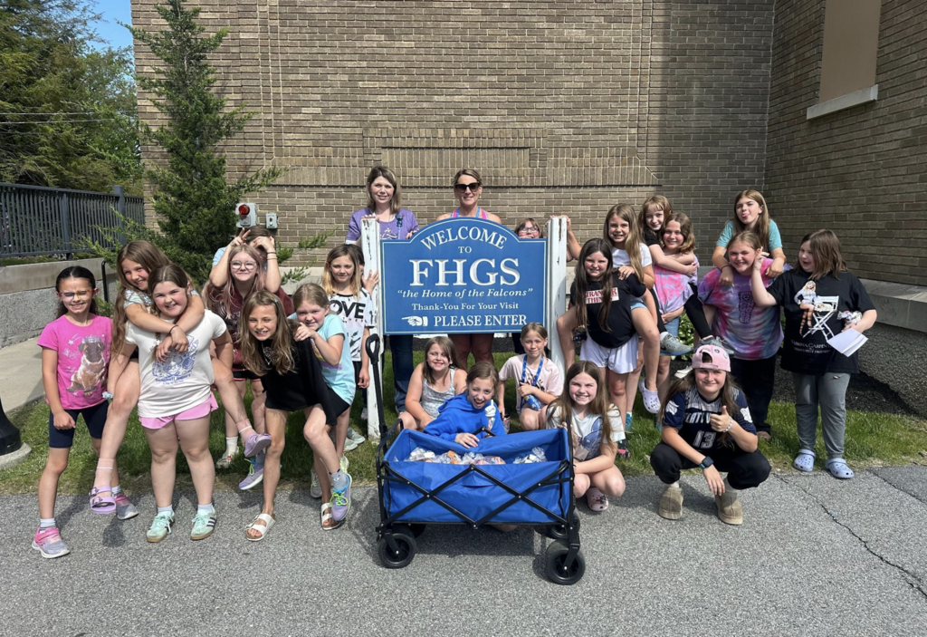 A group of students and staff members gathers around the Fair Haven Grade School welcome sign, posing with a wagon full of donated treat bags.