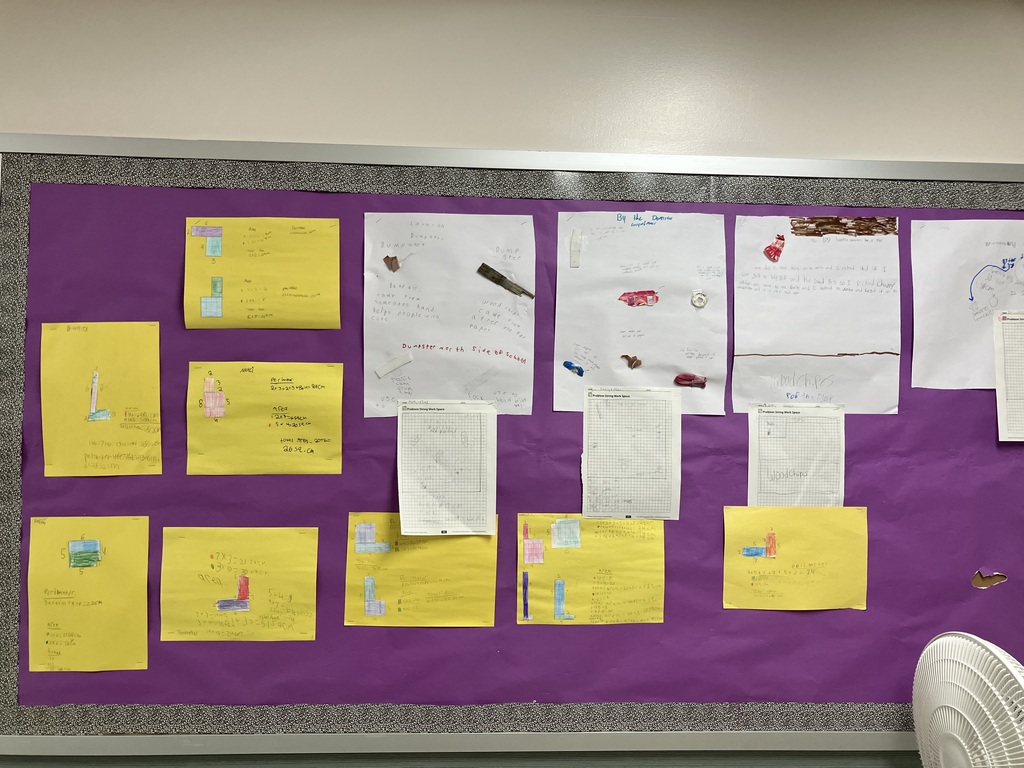 A wide shot of a classroom bulletin board covered in bright purple paper. The board displays a collection of student schoolwork organized into columns. The top row features white poster boards where students have taped found outdoor objects (like wrappers, wood chips, and plastic) alongside written explanations and grid-paper maps. Directly beneath each white poster is a corresponding bright yellow sheet of construction paper. These yellow sheets show colorful, hand-drawn geometric shapes made of square units, paired with student calculations for area and perimeter. A white electric fan is partially visible in the bottom right corner. 