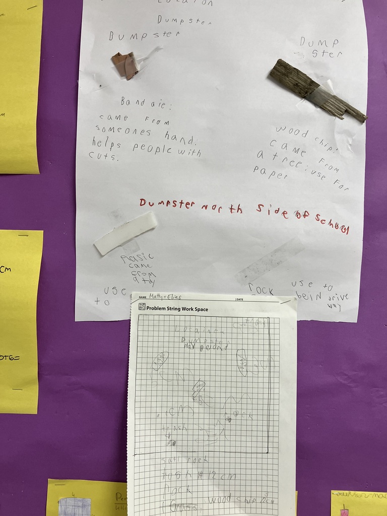  A wide shot of a classroom bulletin board covered in bright purple paper. The board displays a collection of student schoolwork organized into columns. The top row features white poster boards where students have taped found outdoor objects (like wrappers, wood chips, and plastic) alongside written explanations and grid-paper maps. Directly beneath each white poster is a corresponding bright yellow sheet of construction paper. These yellow sheets show colorful, hand-drawn geometric shapes made of square units, paired with student calculations for area and perimeter. A white electric fan is partially visible in the bottom right corner.  