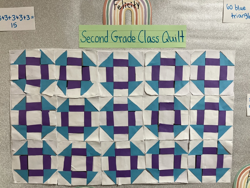  close-up view of the "Second Grade Class Quilt" grid pinned to a glittery silver bulletin board background. The quilt consists of 15 paper squares arranged perfectly in 3 rows and 5 columns. Each individual quilt square has a symmetrical geometric layout: a large white square in the exact center, flanked by four purple rectangles forming an inner ring, and corner elements made of alternating bright blue and white right triangles. The uniform repetition of the shapes across the blocks creates secondary geometric patterns and paths across the rows.