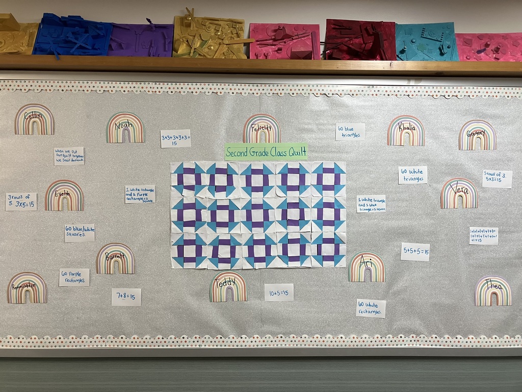  A classroom bulletin board with a silver glitter background displays a math and art project titled "Second Grade Class Quilt" on a green paper banner. In the center is a paper quilt made of 15 individual square blocks arranged in a grid of 3 rows and 5 columns. Each block features a geometric pattern made of white, purple, and bright blue paper shapes. Surrounding the quilt are several boho-style rainbow cutouts labeled with students' names (such as Collin, Noah, Evelyn, Barrett, Remmington, Teddy, Felicity, Khalia, Gregory, Vera, Ari, and Thea). Next to each name is a small handwritten note containing math equations summing to 15 or calculations counting the different colored shapes used across the entire quilt.  