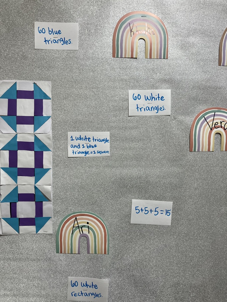 A close-up view of the "Second Grade Class Quilt" grid pinned to a glittery silver bulletin board background. The quilt consists of 15 paper squares arranged perfectly in 3 rows and 5 columns. Each individual quilt square has a symmetrical geometric layout: a large white square in the exact center, flanked by four purple rectangles forming an inner ring, and corner elements made of alternating bright blue and white right triangles. The uniform repetition of the shapes across the blocks creates secondary geometric patterns and paths across the rows. 