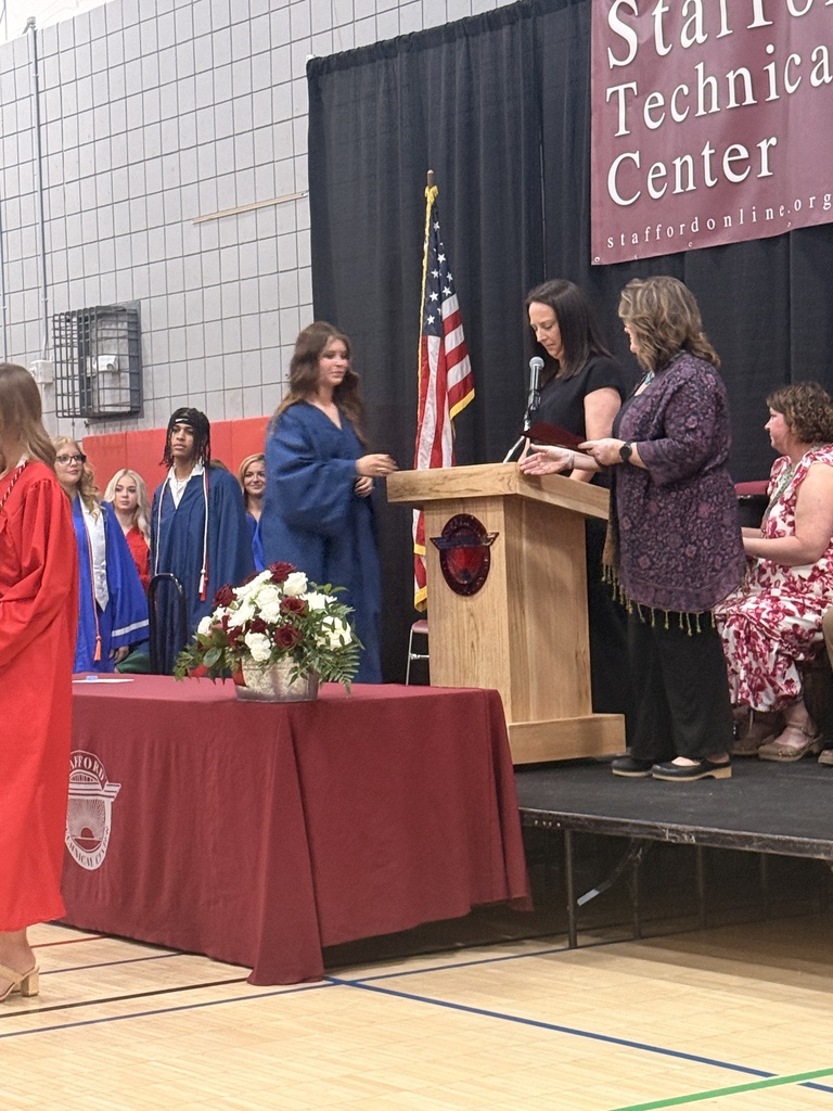 A student in a blue graduation gown approaches the podium to receive recognition. Other graduates and staff are visible on and near the stage.