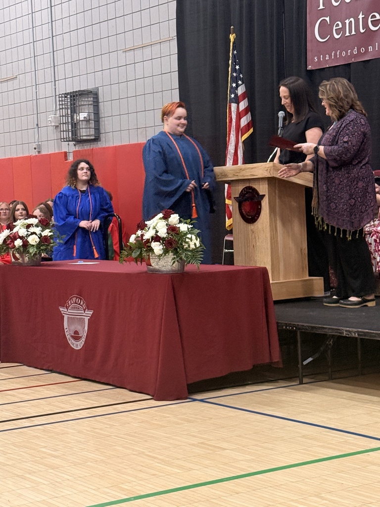 A student in a blue graduation gown and honor cords approaches the podium during the Stafford Technical Center awards ceremony while classmates watch from their seats.