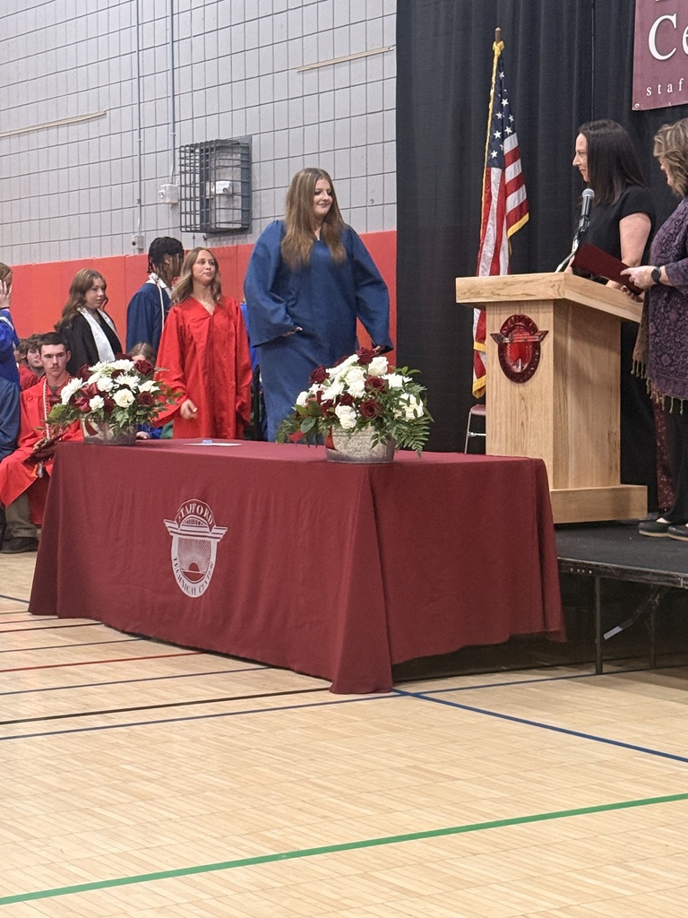 A student in a blue graduation gown smiles while walking toward the stage as fellow graduates wait in line behind her during the awards ceremony.
