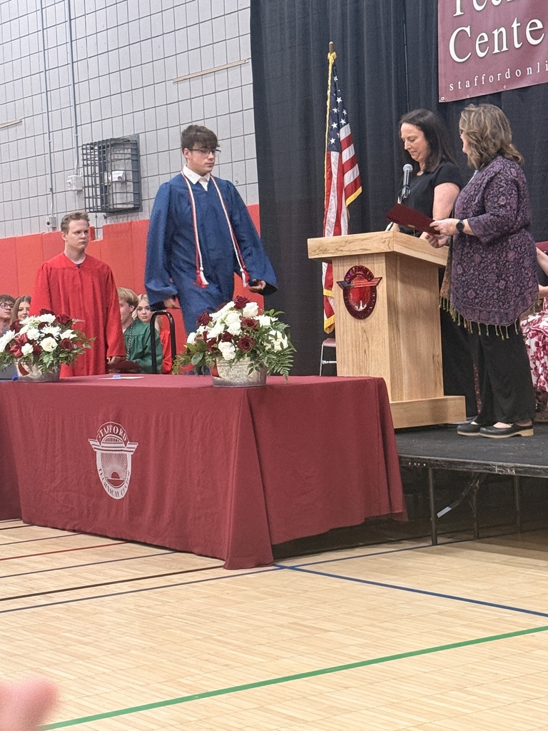 A student in a blue graduation gown approaches the stage to receive recognition while two presenters stand at a podium. Other graduates wait nearby.