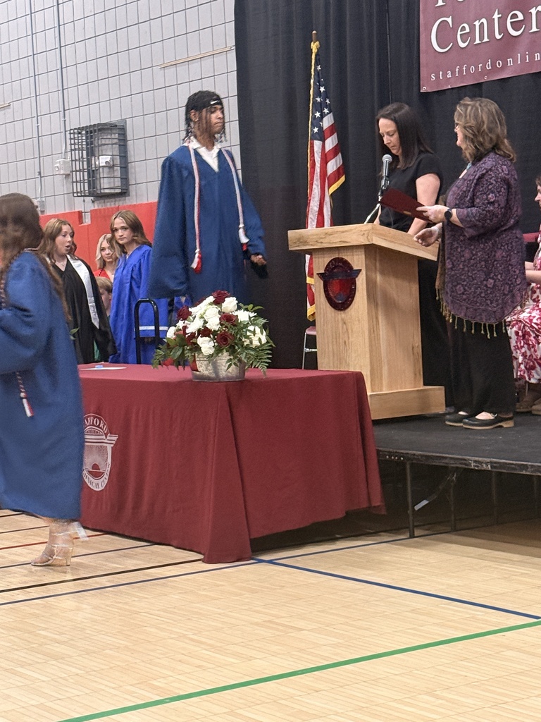 A student in a blue graduation gown with honor cords walks toward the stage while presenters stand at the podium and graduates wait nearby.