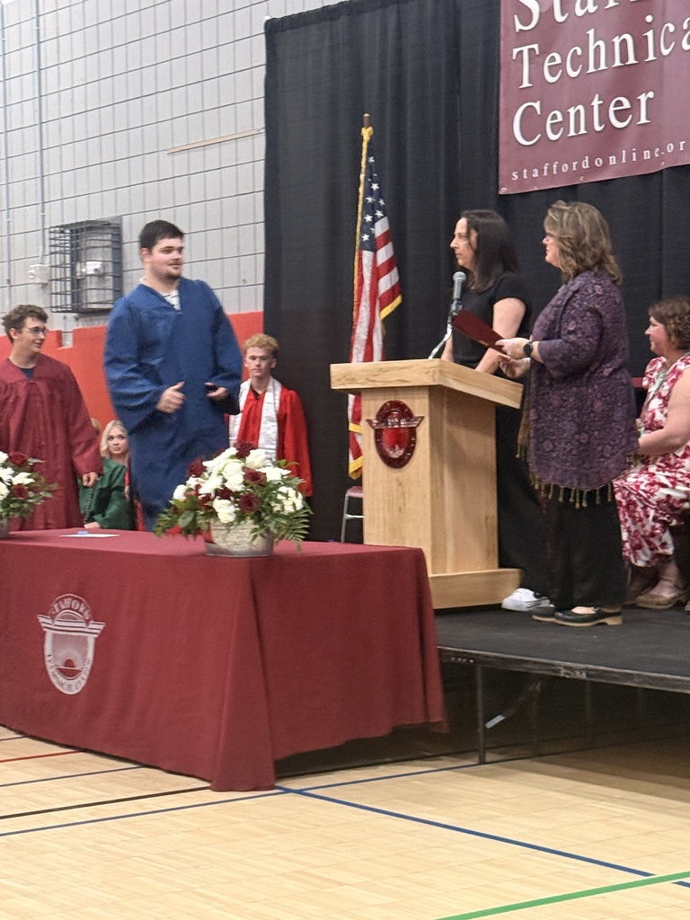 A student in a blue graduation gown approaches the stage as presenters stand at the podium. Graduates seated nearby watch the ceremony.