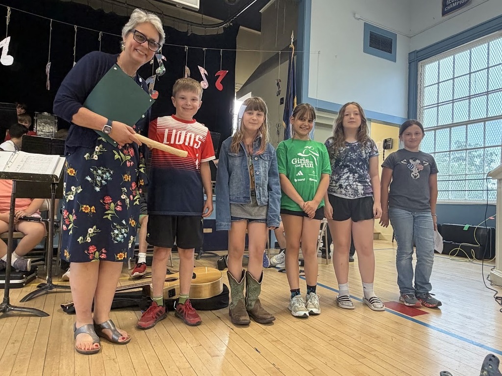 Mrs. Rampone stands smiling with a certificate at an Orwell Village School PBIS assembly. She is joined by five elementary students standing in a row beside her on the gymnasium floor. Musical instruments and student performers are visible in the background.