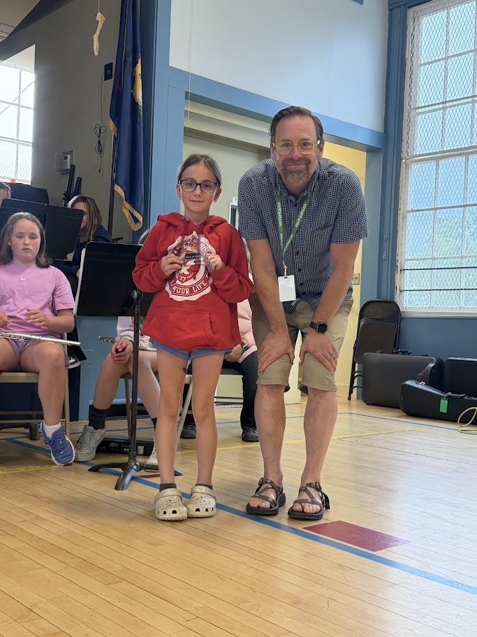 A student wearing a red sweatshirt stands with a teacher during an award presentation at an Orwell Village School assembly. The student holds a small prize while classmates sit behind them.
