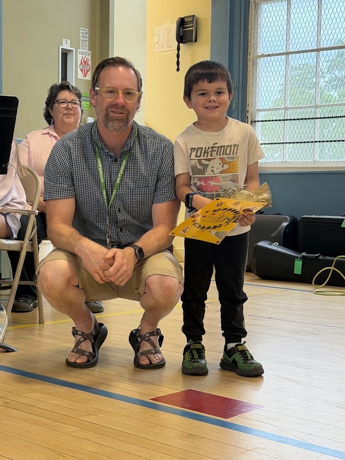 A teacher kneels beside a smiling student holding a certificate and award during an Orwell Village School assembly. A woman is seated in the background while musical equipment and student performers are visible nearby.