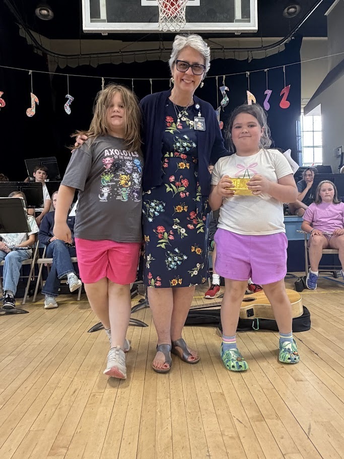Mrs. Rampone stands smiling between two students during an awards presentation at Orwell Village School. One student holds a small gold-colored award while classmates and musical instruments are visible in the background.